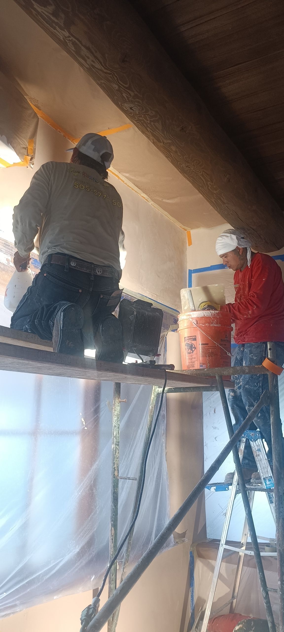 Two workers on scaffolding install ceiling panels in a room under construction.