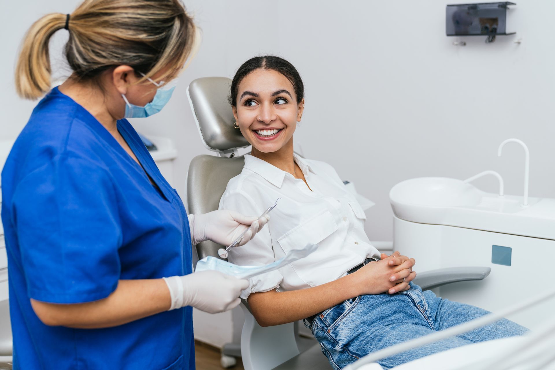 A cosmetic dentist with blue clothing is attending to a smiling young woman in her dental office.