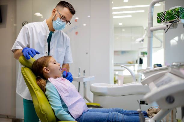 A young girl is seated in a dentist chair with a man behind her, highlighting a family dentist.