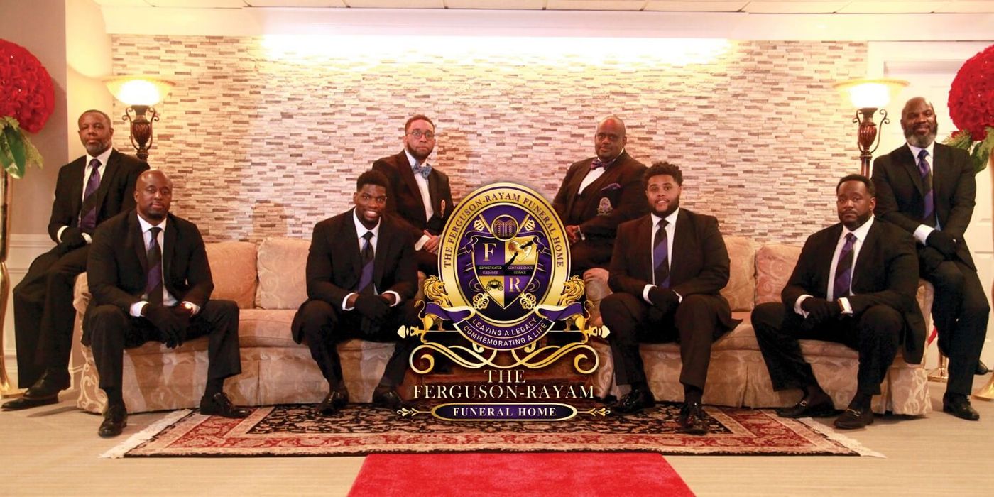 Group of people in dark suits posing indoors behind a gold emblem on a brick wall.