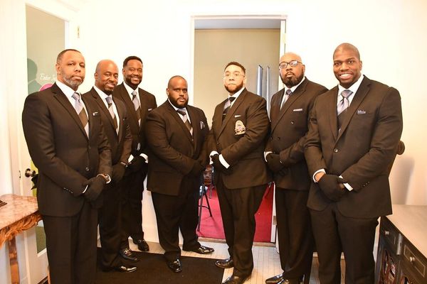 Six men in dark suits standing indoors in a hallway, posing for a group photo.
