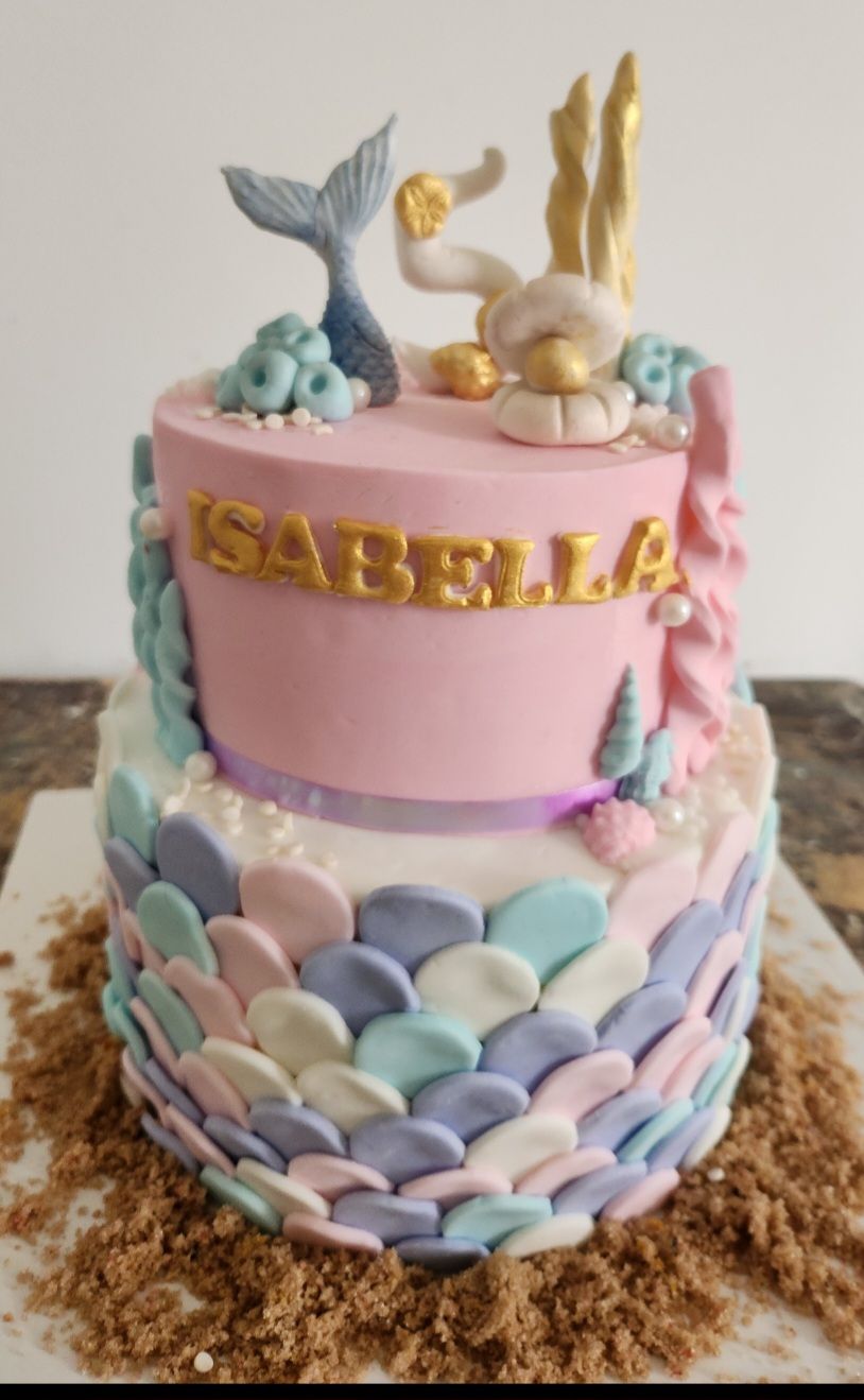 Pastel de dos niveles con temática de sirena con glaseado rosa, cola de sirena, detalles dorados y nombre 