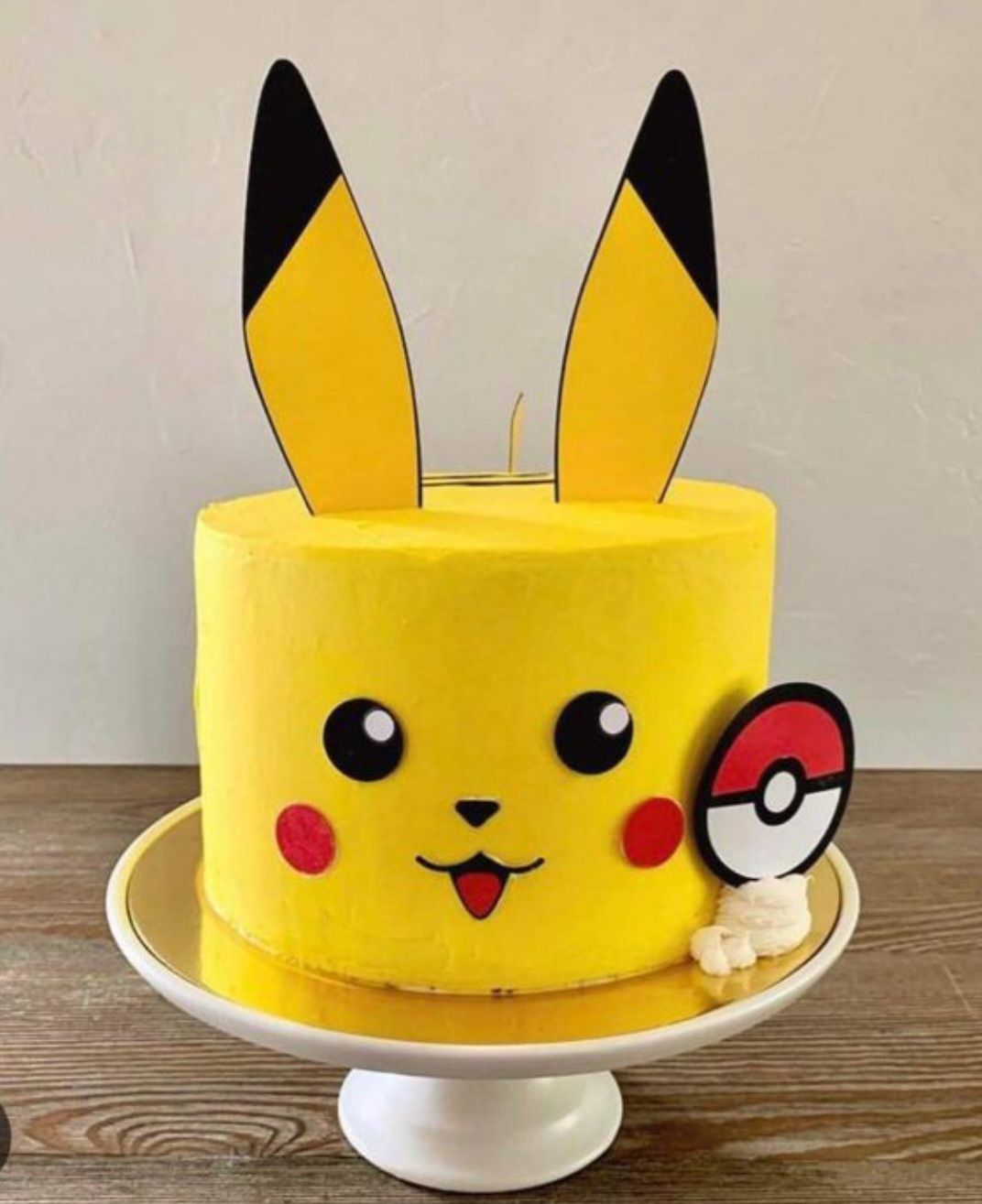Pastel con temática de Pikachu con glaseado amarillo, orejas negras y una Pokébola sobre un soporte para pastel blanco.