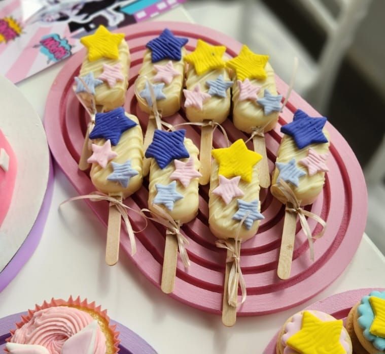 Cake pops cubiertos de chocolate blanco, con palitos y decorados con estrellas. Servidos en una bandeja rosa.