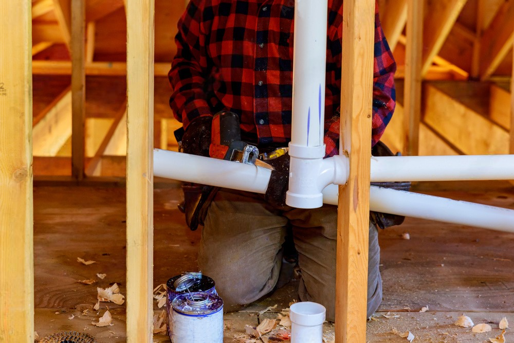 Plumber working on white PVC pipes inside a wooden-framed house under construction