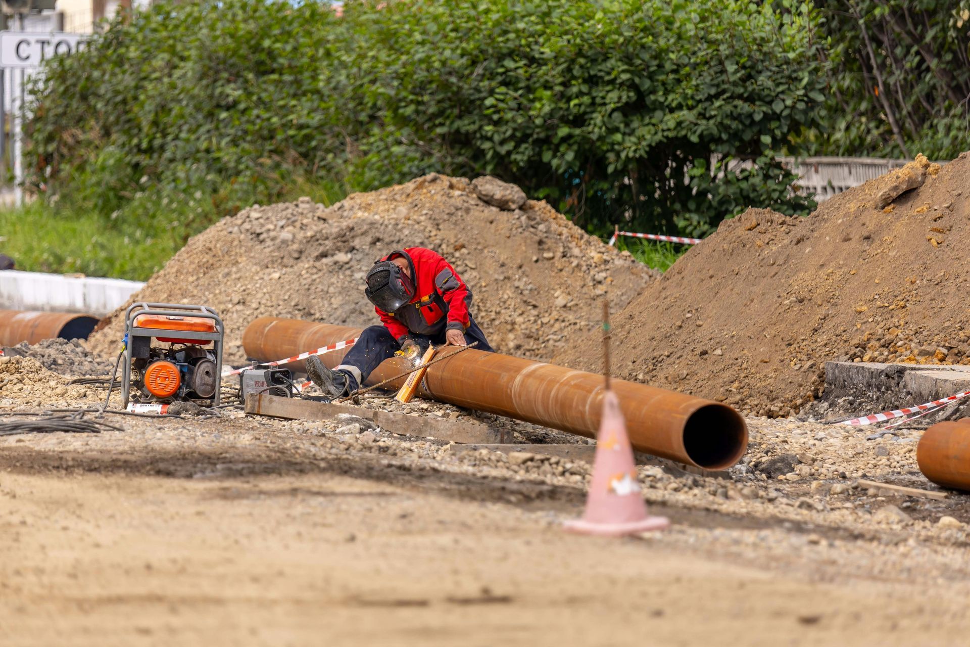 Worker in red jacket cutting a large pipe at a road construction site near dirt piles and an orange machine