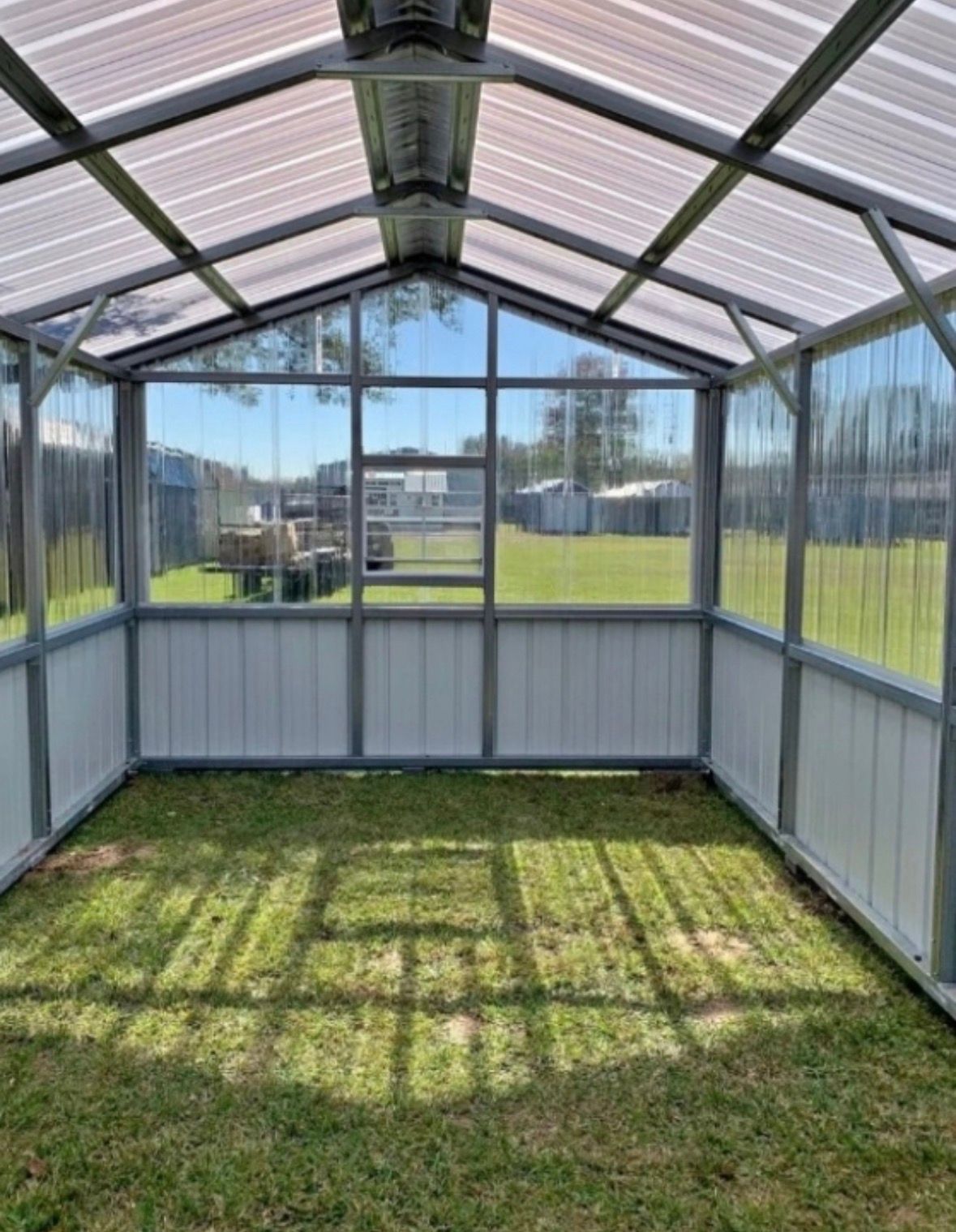 Empty greenhouse with a metal frame, glass roof, and grassy floor under bright sunlight