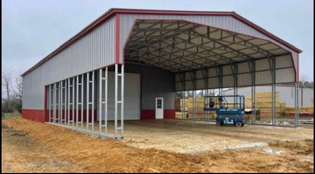 Large metal barn under construction with open front, gray siding, and a blue lift on a dirt lot.