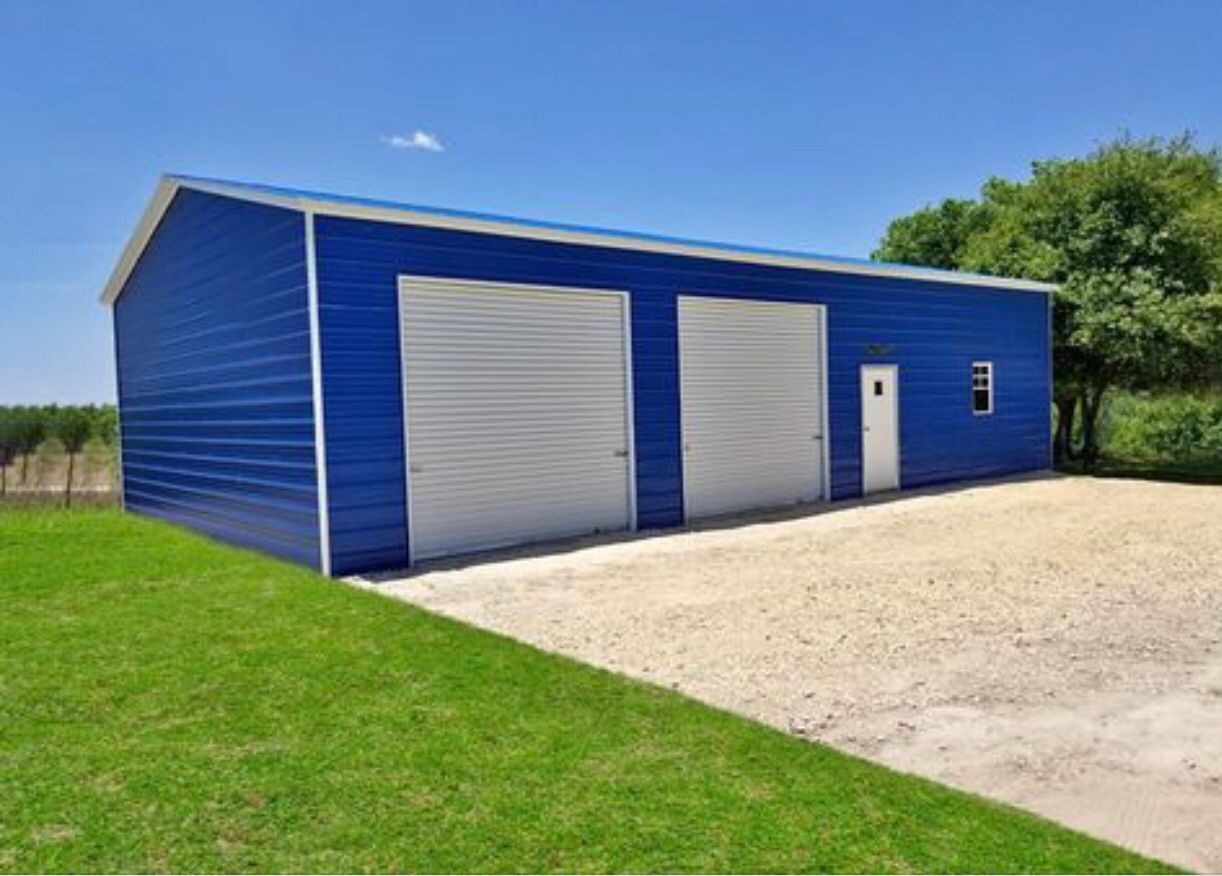 Blue metal storage building with white doors beside a gravel lot and grassy field
