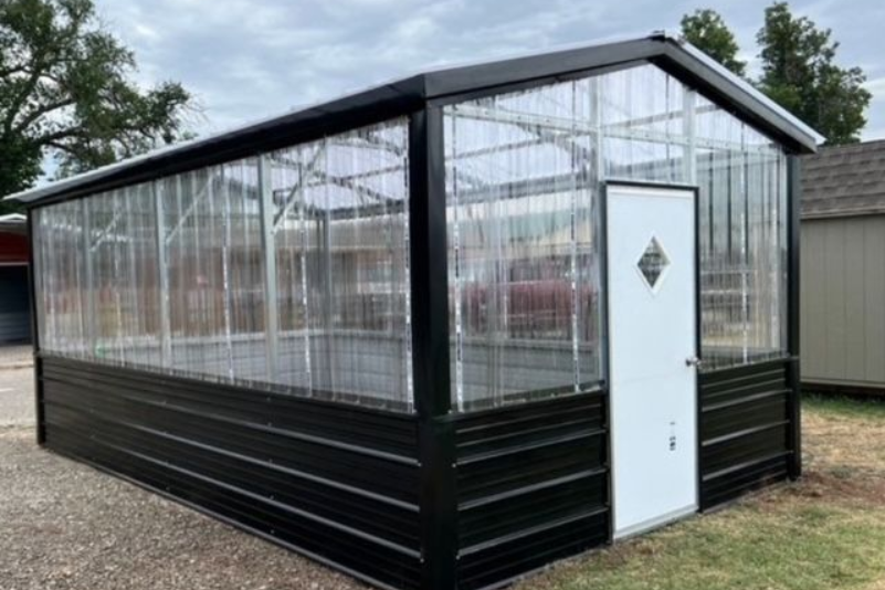 Black-framed glass greenhouse with a white door on a gravel lot