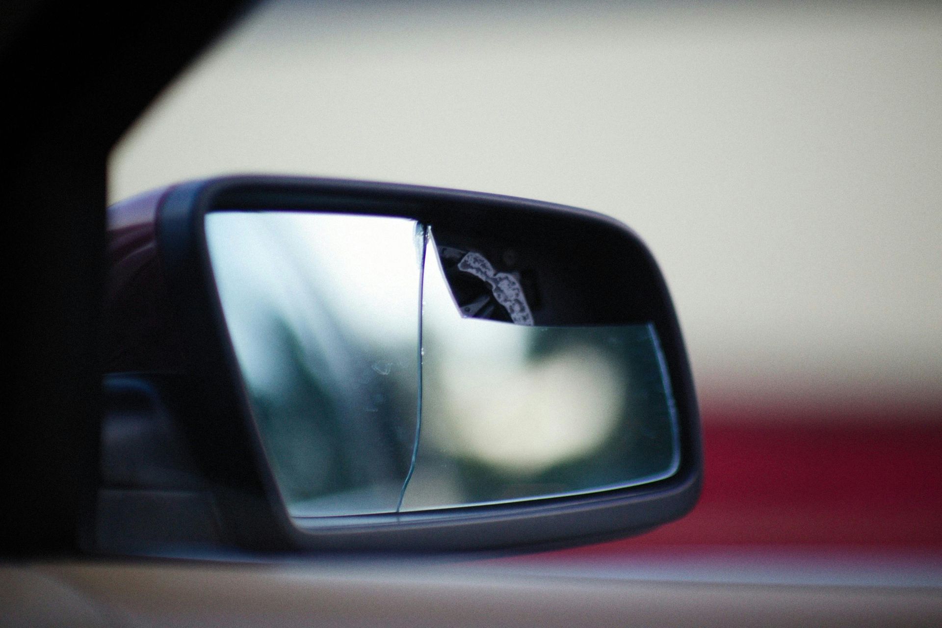 Side mirror of a car reflecting a blurred road and sky, with a red vehicle edge in the foreground