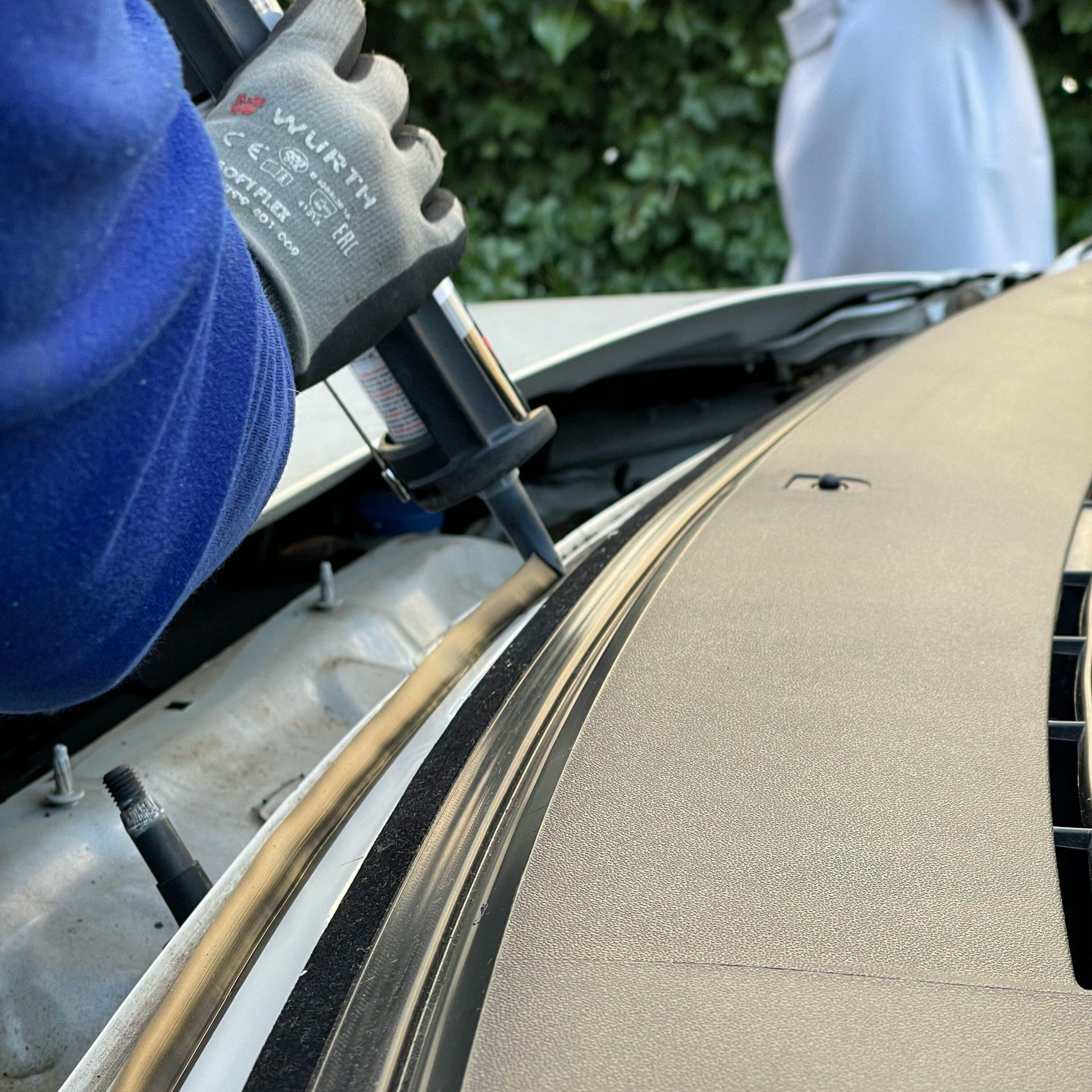 Gloved worker applying adhesive along a car windshield edge outdoors