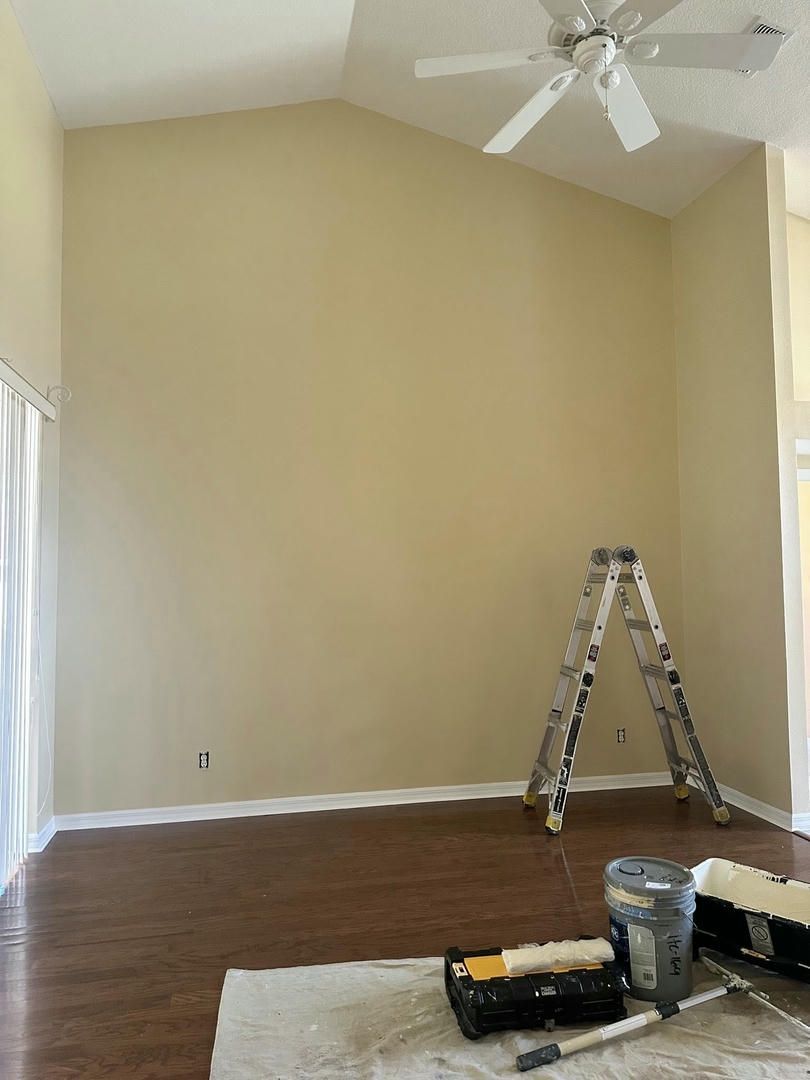 Tall beige wall with a stepladder and painting supplies in a room with hardwood floors.
