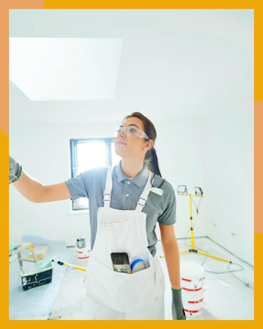 Woman wearing safety glasses and paint-stained overalls paints a white room with a skylight.