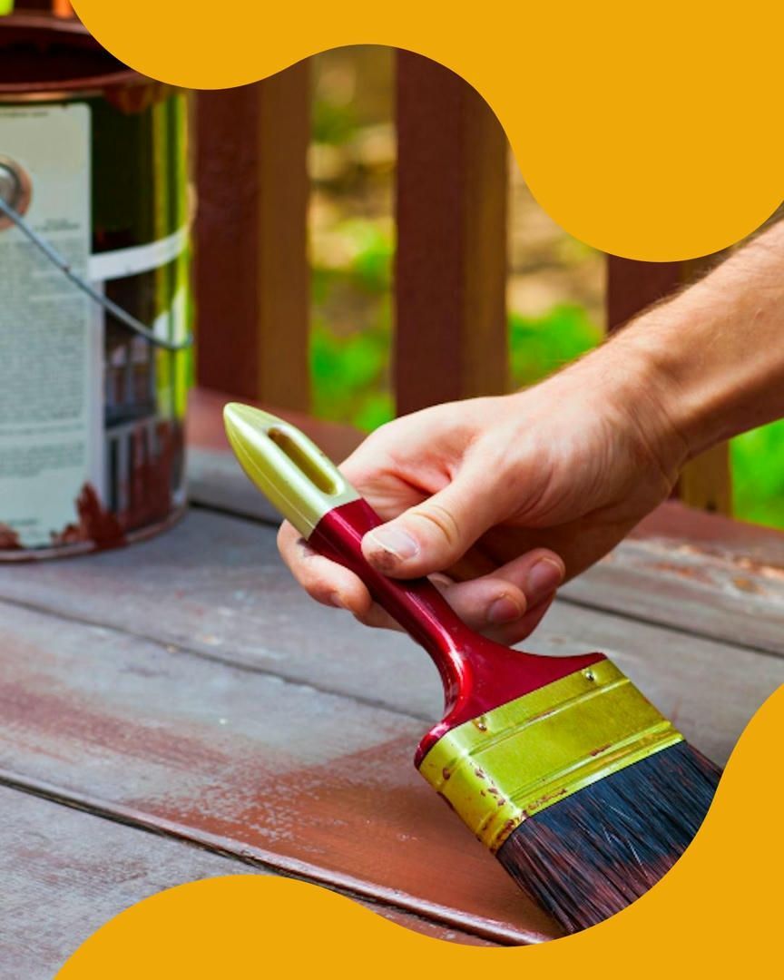 Person's hand holds paintbrush, painting a wooden deck brown, paint can nearby.