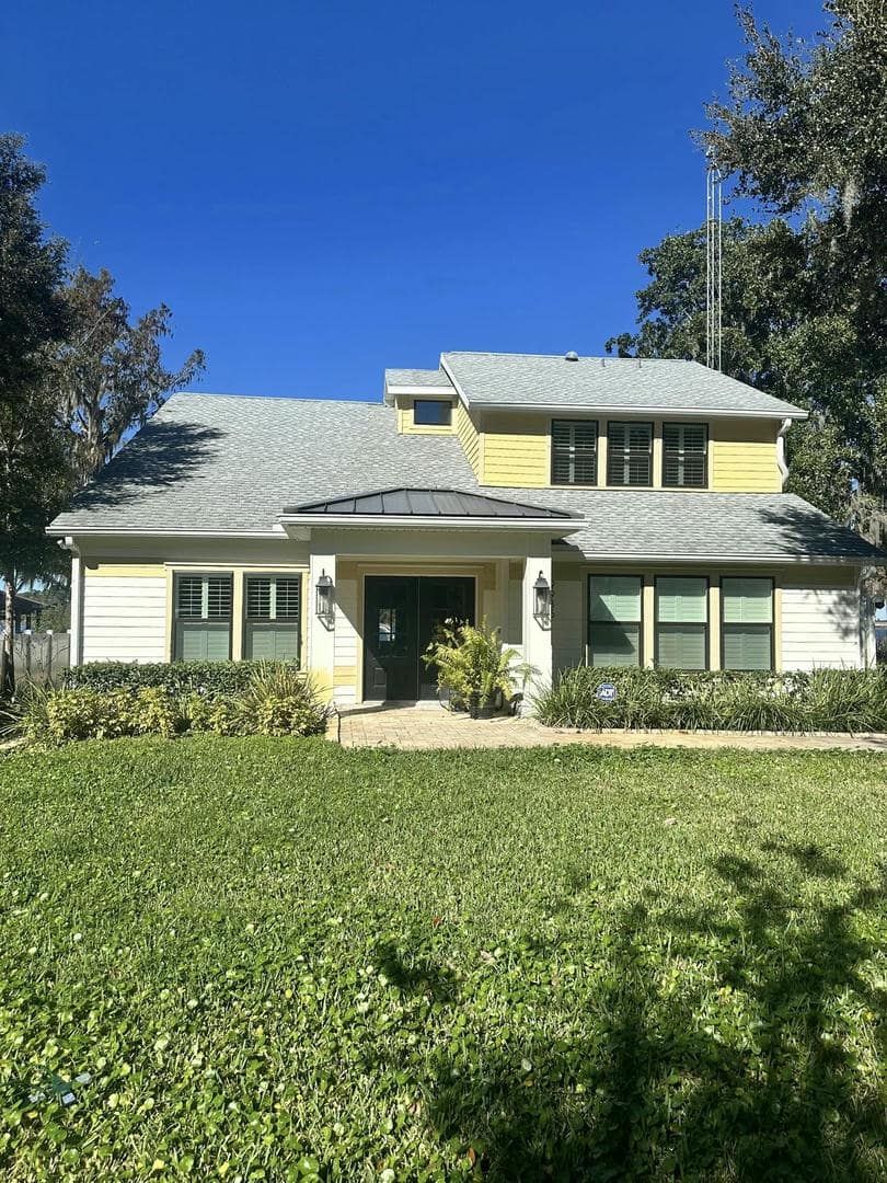 House with light yellow siding, gray roof, and green lawn under a blue sky.