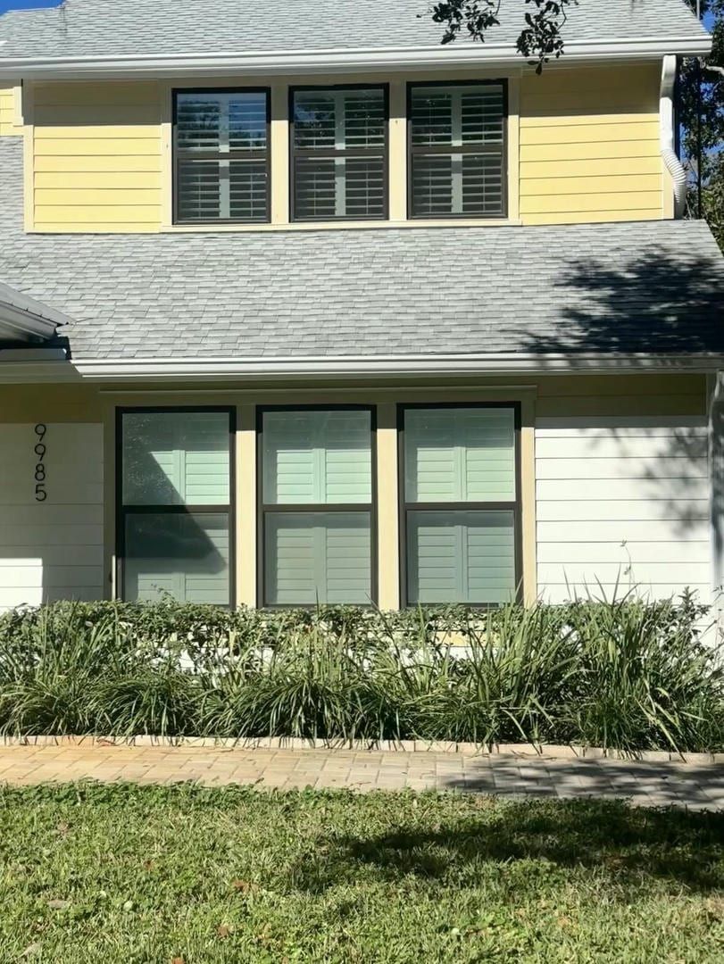House with light yellow siding, dark-framed windows, and a gray roof. Green shrubs line the front.