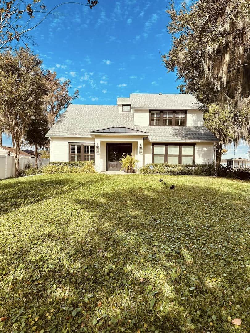 House with beige siding, dark trim, and gray roof, situated on a grassy hill with blue sky.