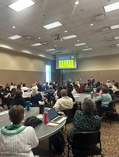 Large conference room; people seated at tables, listening to a presentation on a screen.