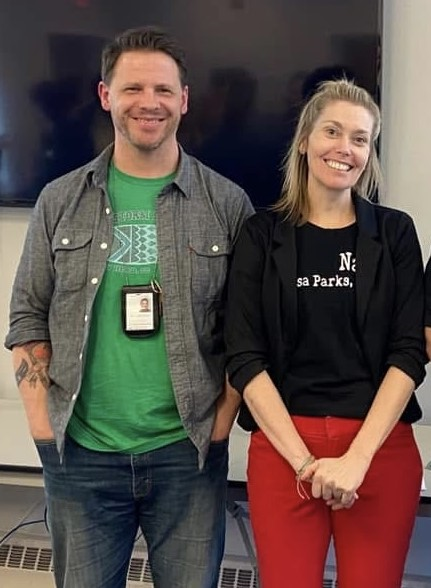 Man in gray shirt and jeans, and woman in black blazer and red pants, smiling. Standing inside.