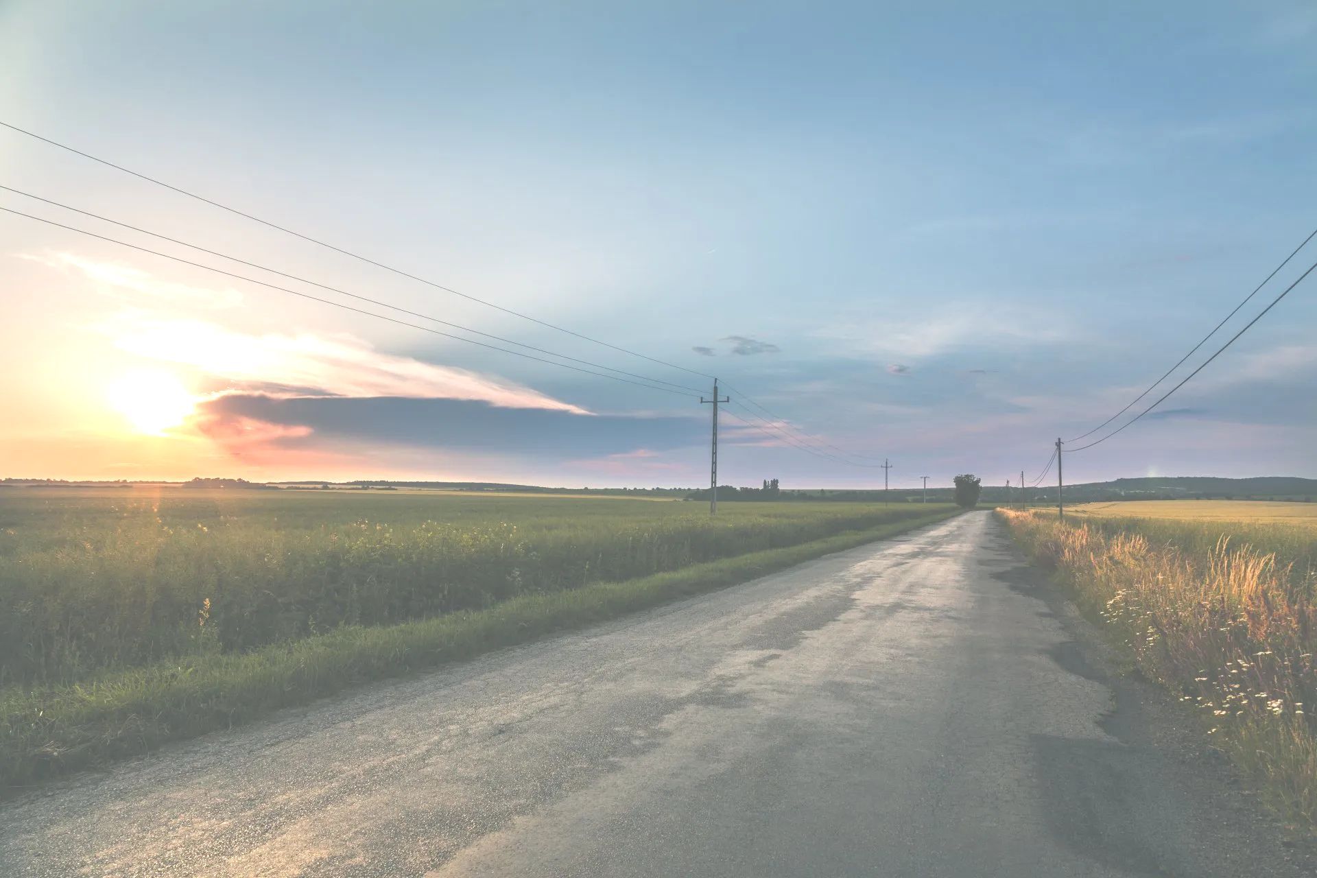 Rural road at sunset, flanked by fields, with power lines overhead. Sky is blue with orange and pink hues.