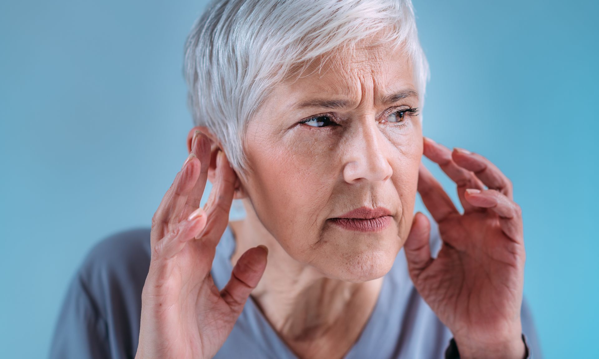 An elderly woman is covering her ears with her hands.