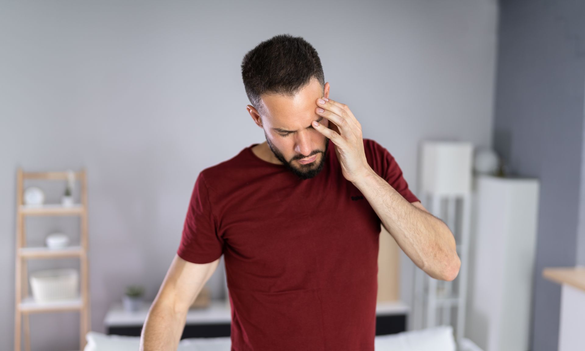 A man is holding his head in pain while standing in front of a bed.