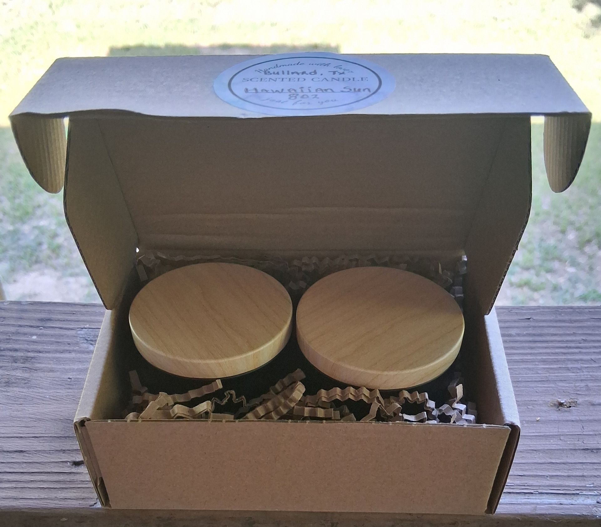 Open cardboard gift box with two round wooden lids and packing filler inside on a table.
