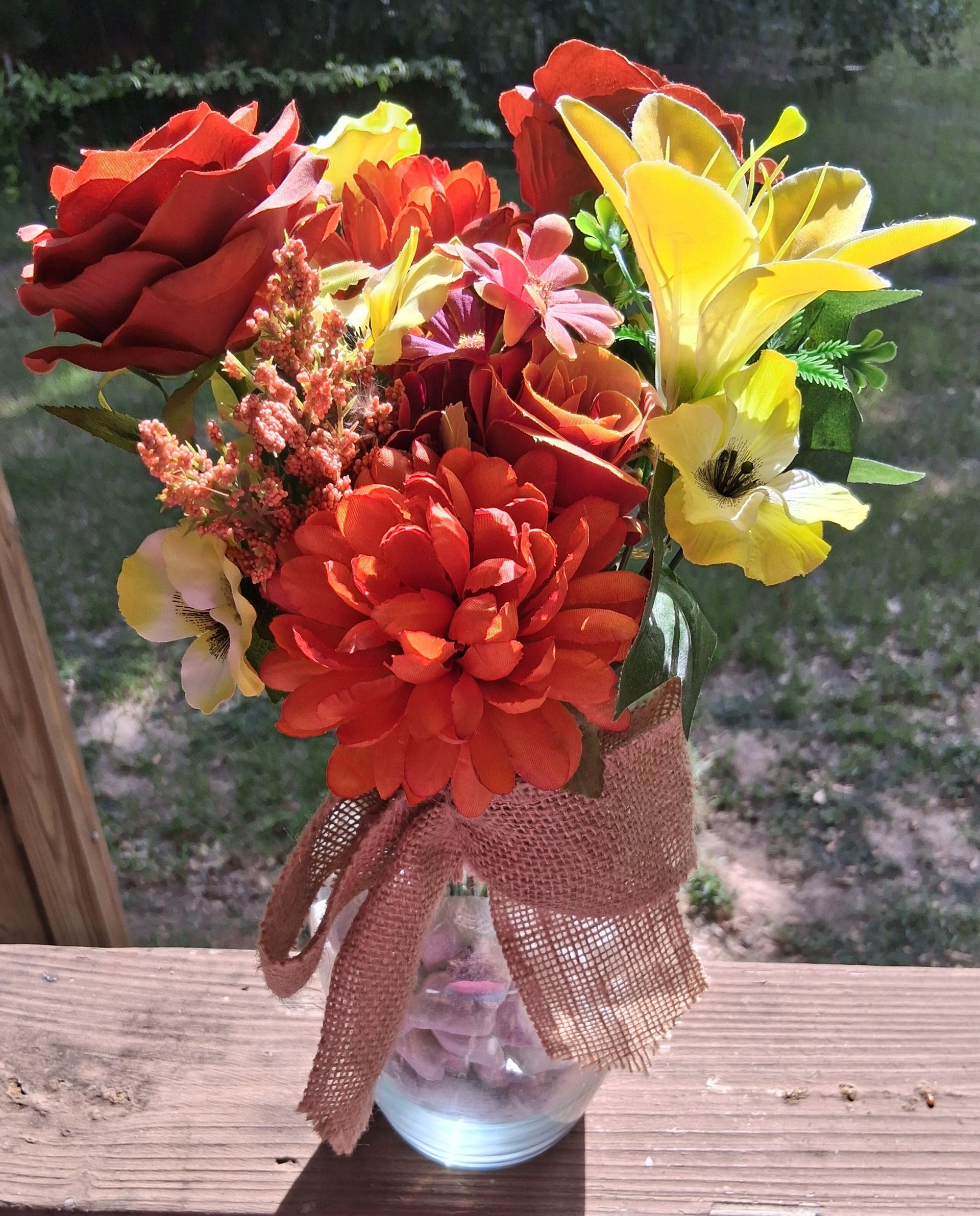 Bouquet of purple, yellow, and white flowers in a glass vase with a burlap bow on a wooden table outdoors