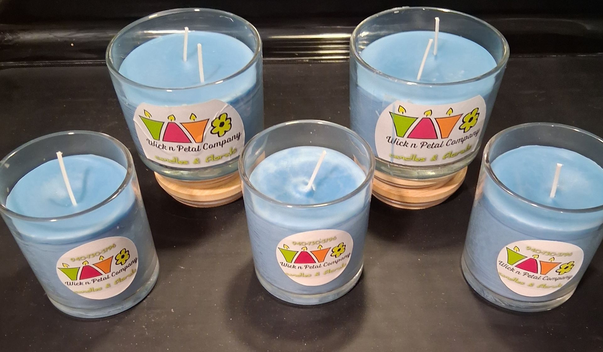 Five blue candles of varying sizes arranged on a dark surface, each in a clear glass jar with a colorful label