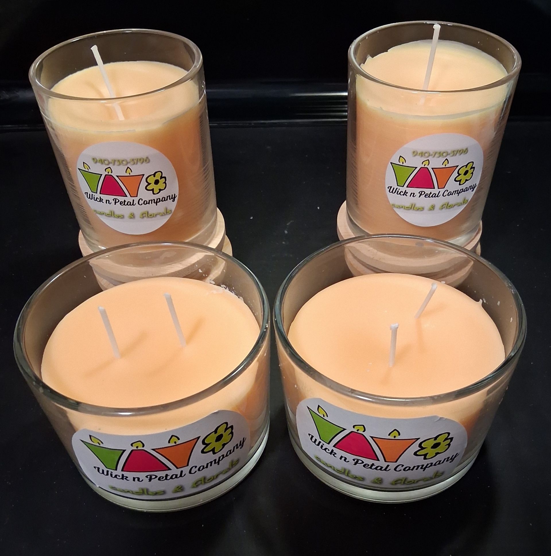 Four peach-colored candles in clear glass jars arranged in a 2x2 grid on a dark surface.