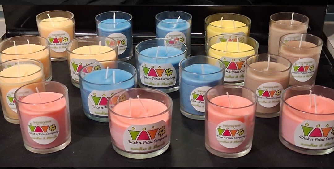 Assorted colorful candles in glass jars arranged on a table, with pink, blue, yellow, and beige labels.