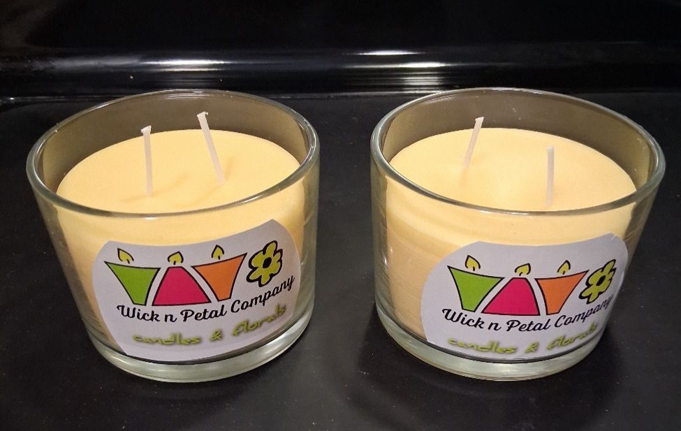 Two cream-colored candles in glass jars on a dark surface with colorful labels.