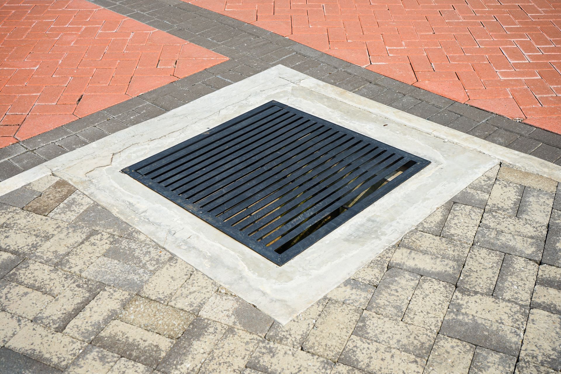 Square street drain grate set in a concrete and brick sidewalk corner.