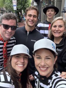 A group of six people posing for a smiling selfie, some wearing striped shirts and hats, outdoors on a city sidewalk.