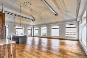 An expansive, modern open-plan loft with hardwood floors, a kitchen island, high vaulted ceilings, and large windows.
