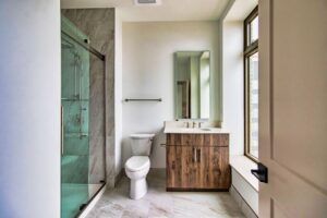 A modern bathroom featuring a wood-toned vanity, a toilet, a glass-enclosed shower, and a window with natural light.