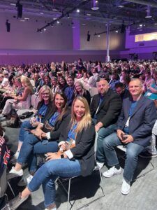 A group of people sitting in chairs in a large conference hall, looking toward the camera.