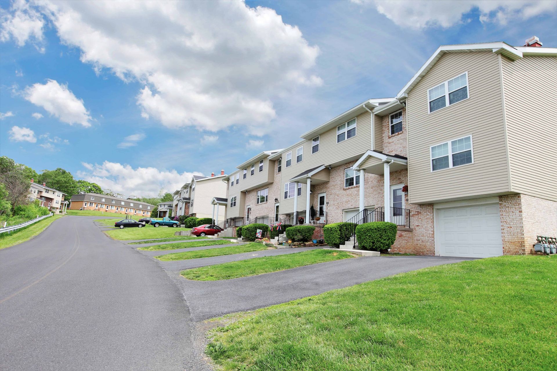 Townhouses along a street, with driveways, grass, and a cloudy sky overhead.