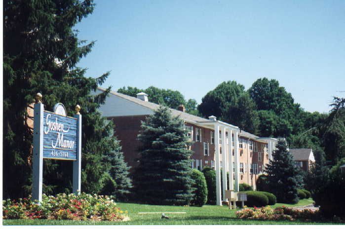 A sign for Goshen Manor apartments stands in front of a multi-story brick apartment building surrounded by trees.