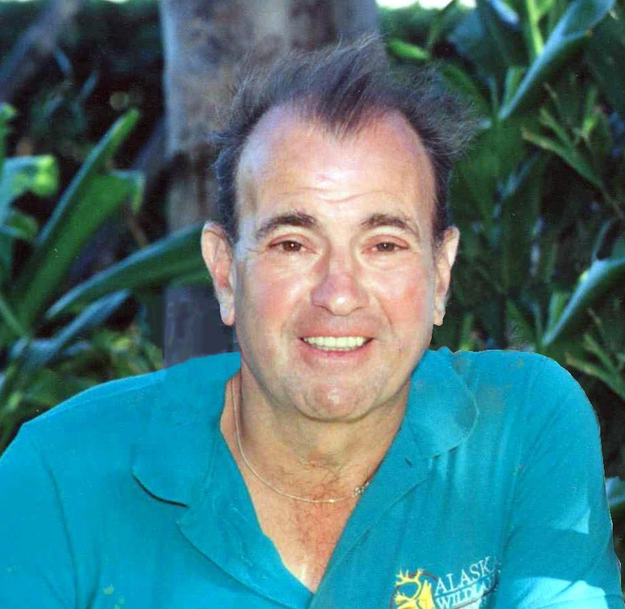 A smiling person wearing a teal shirt outdoors with green foliage in the background.