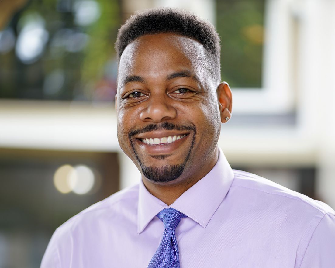 Man in lavender shirt and blue tie smiles, indoors with blurred background.
