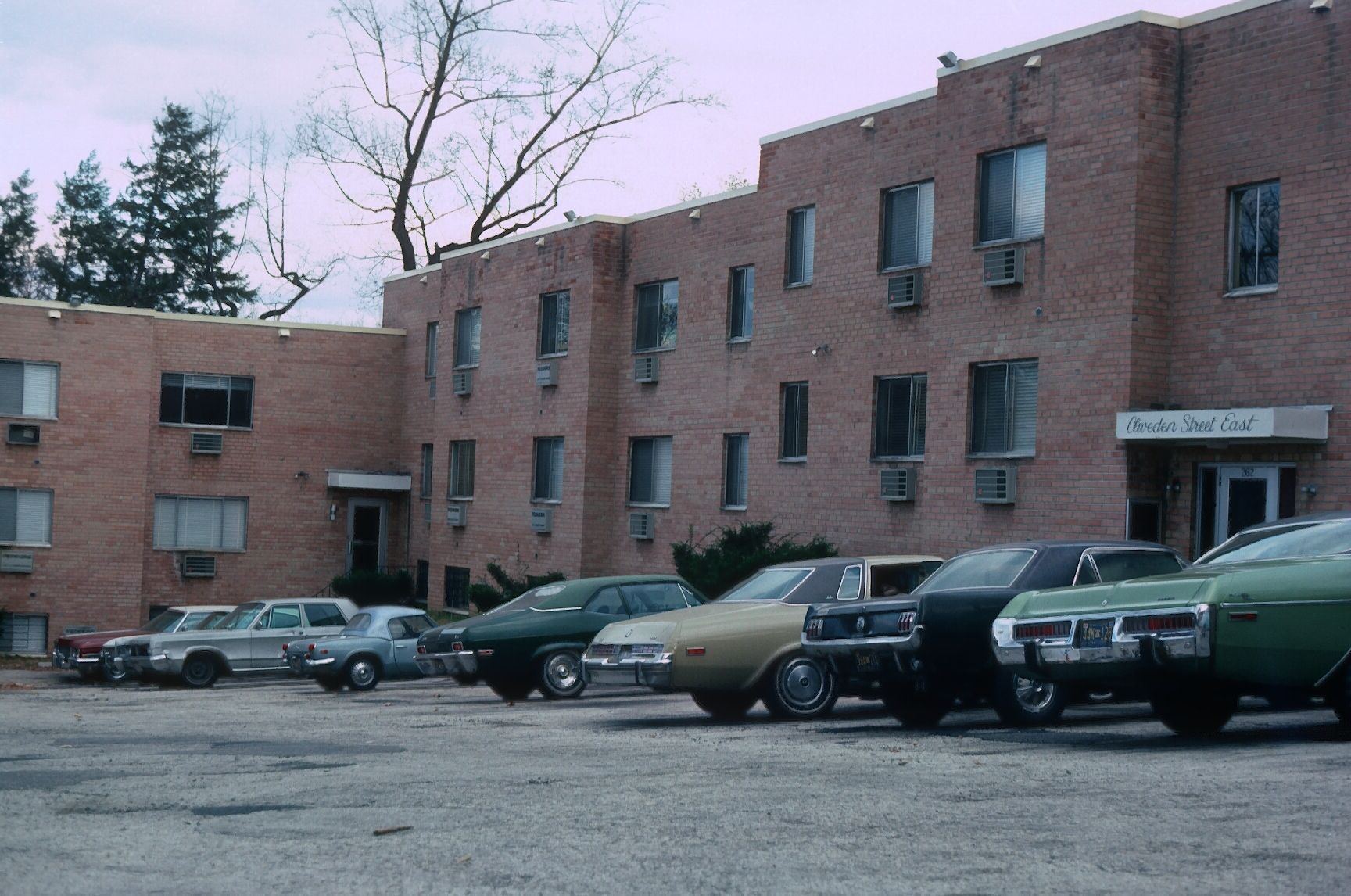 A brick apartment building with a parking lot containing several classic cars in front.