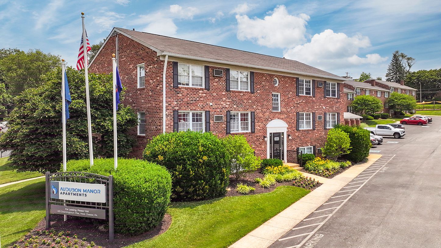 Brick apartment building with white-trimmed windows, American flags, and parked cars along a driveway