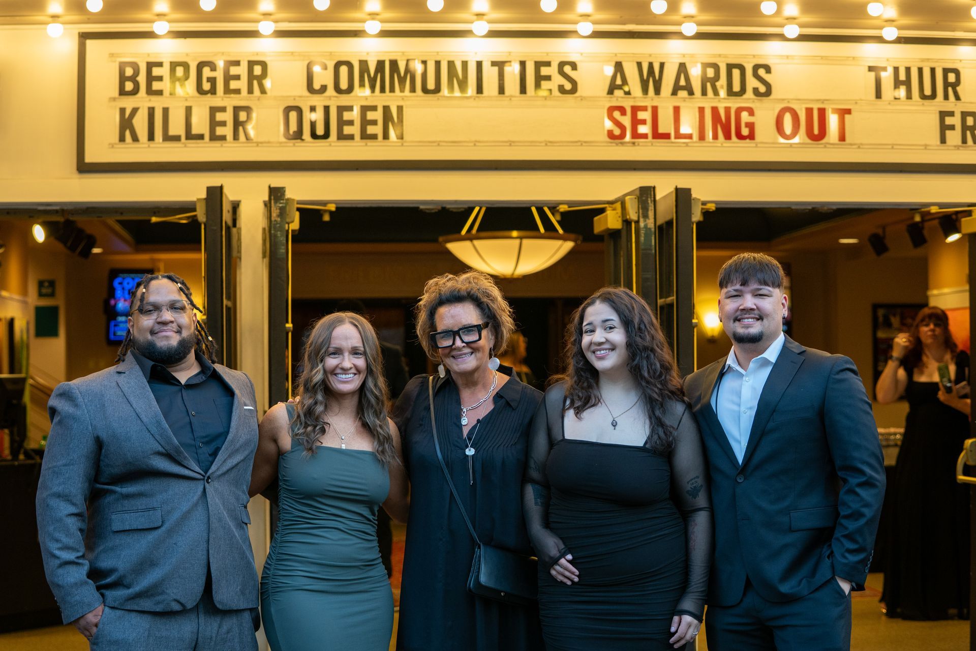 Five people posing outside a theater marquee reading “Killer Queen” and “Selling Out.”
