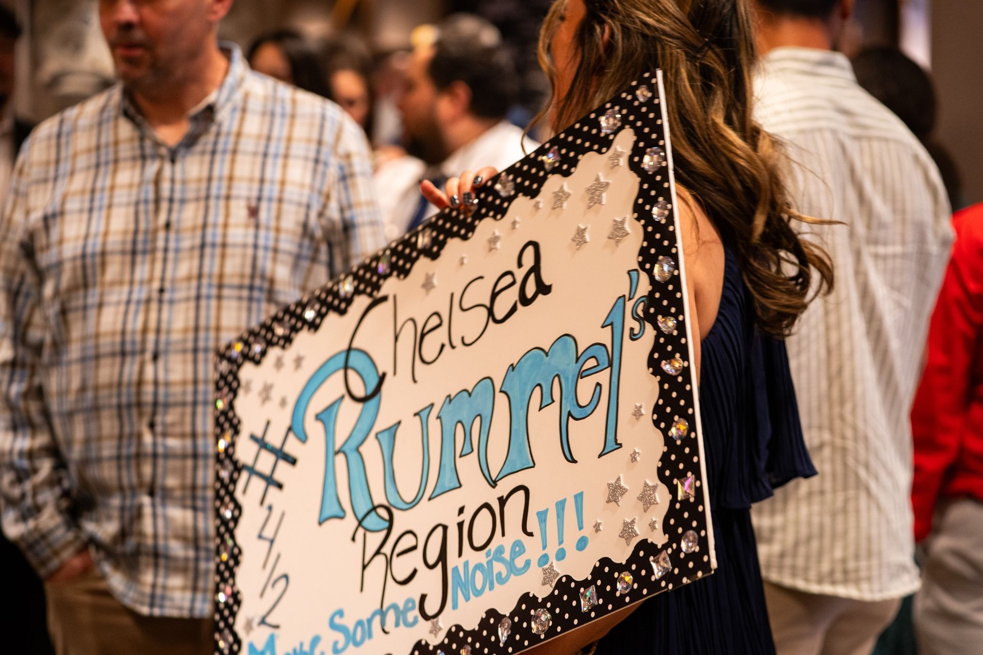 Person holding a decorated sign reading “#Heised Rumpru Region!!!” in a crowded indoor protest scene