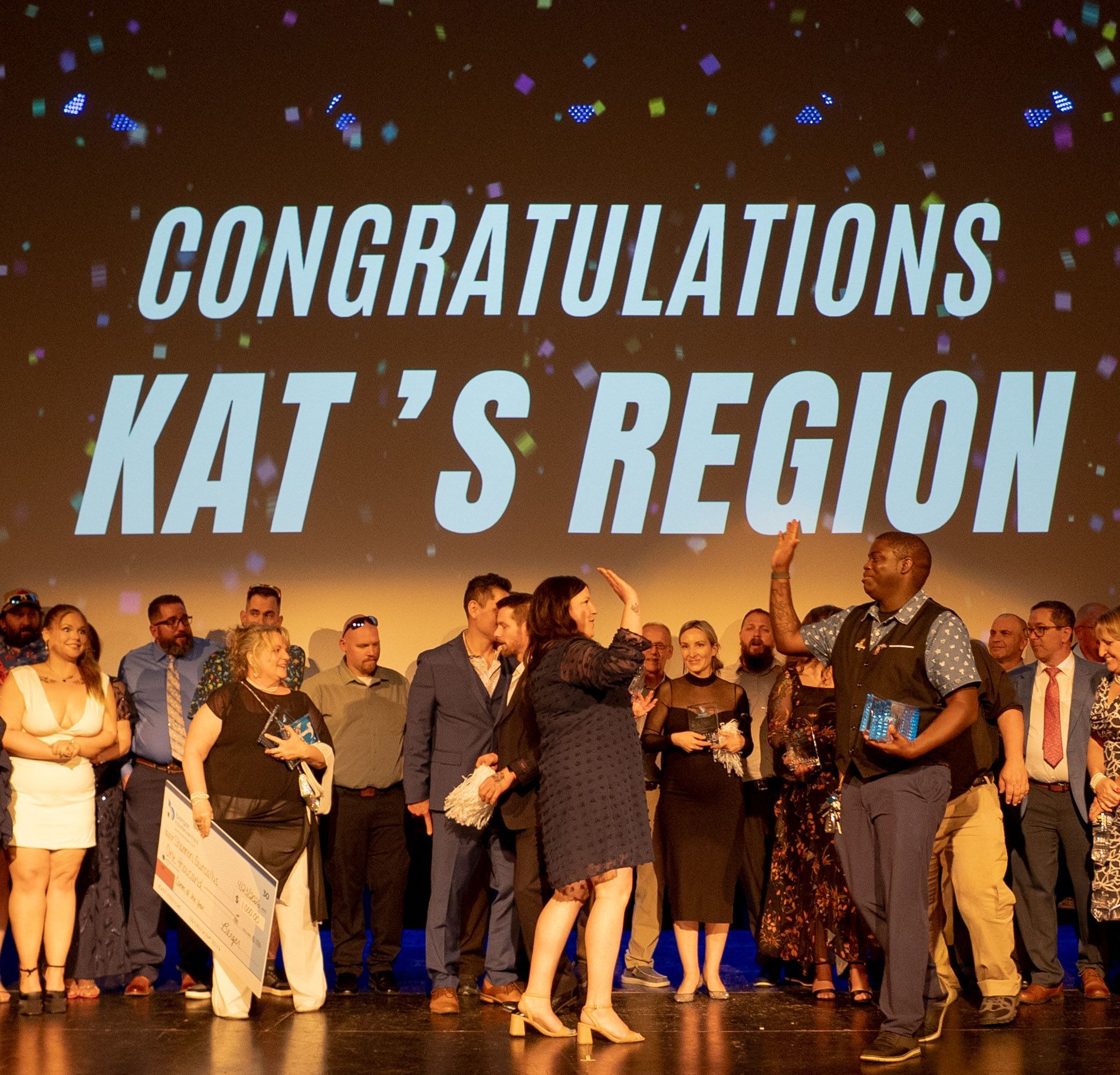 People on stage under “CONGRATULATIONS KAT’S REGION” banner, applauding and celebrating