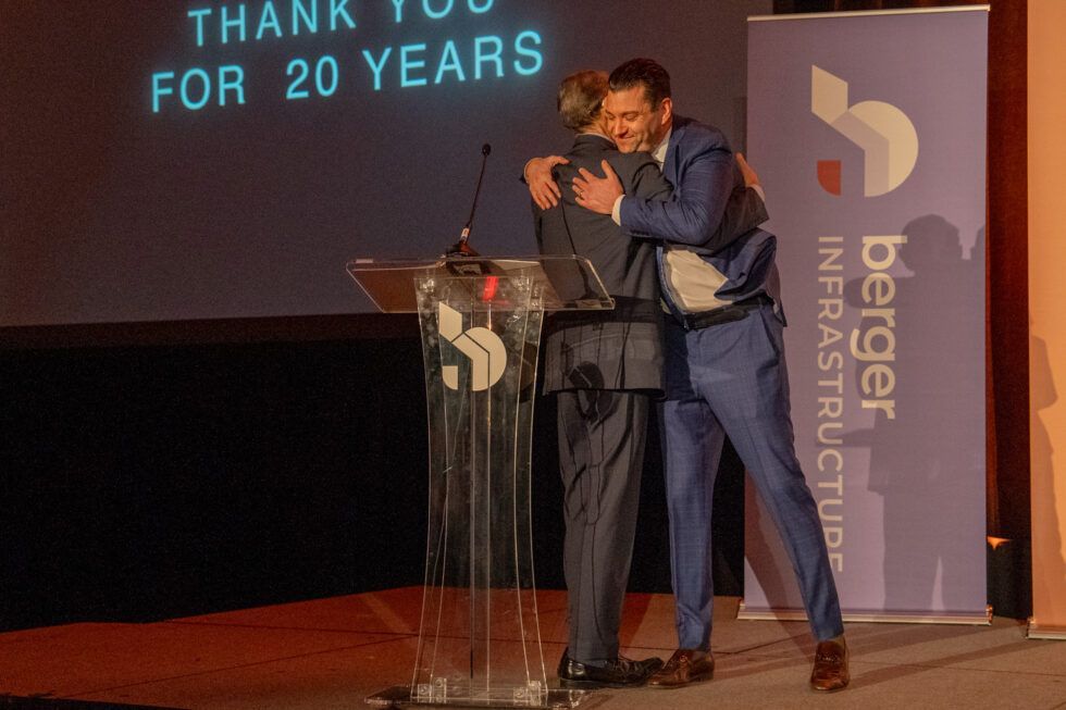Two individuals in suits embrace behind a podium labeled
