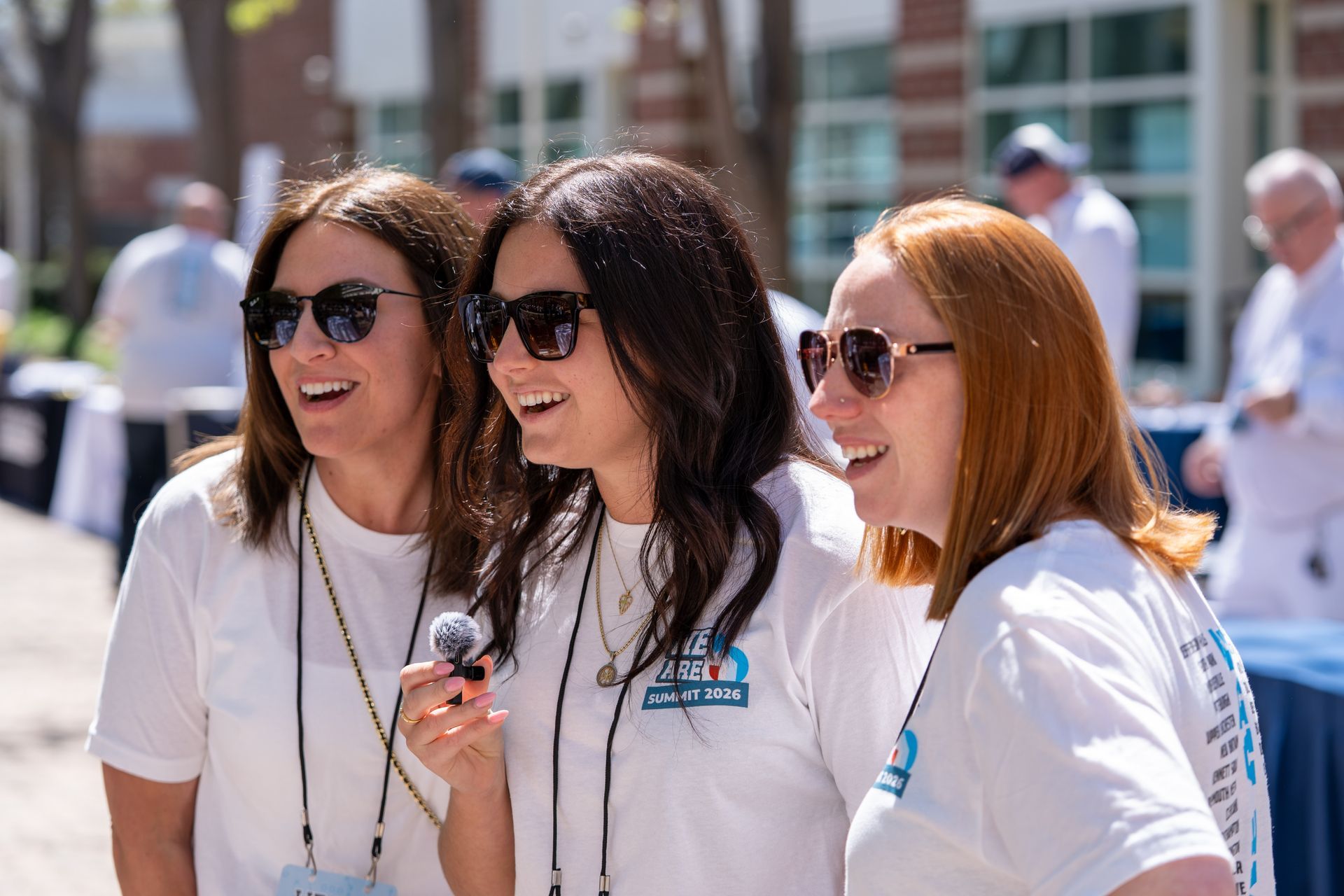Three people in white shirts and sunglasses smiling outdoors, holding a microphone.