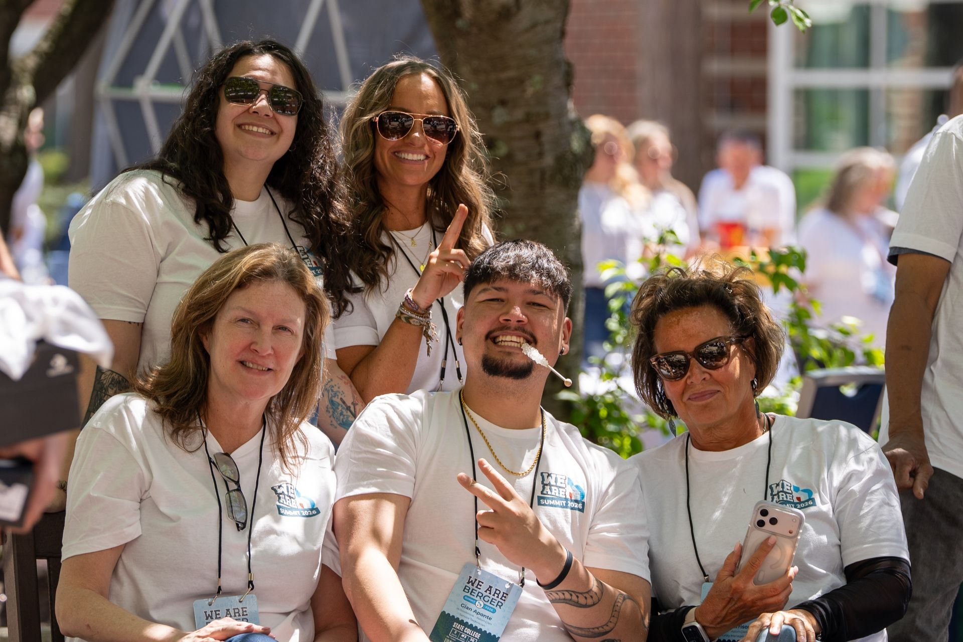 Five smiling people in white shirts posing outdoors at an event, holding lanyards and cups.